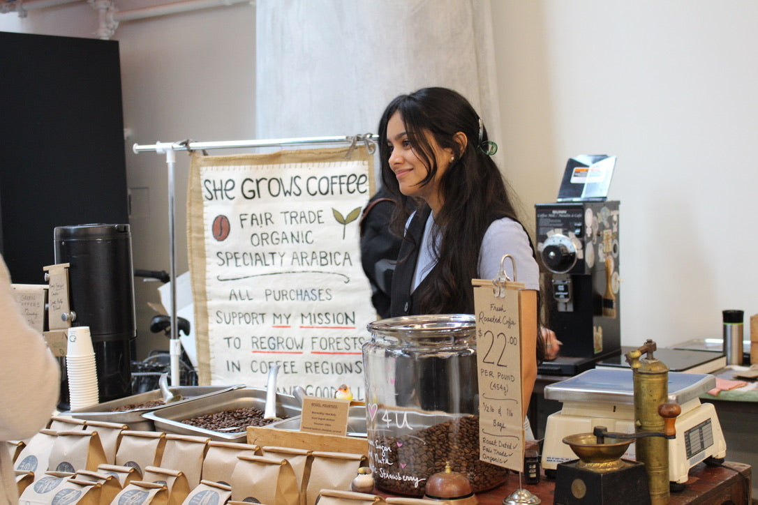 A She Grows Coffee team member at a market booth, with organic beans, vintage equipment, and a sign supporting reforestation. She Grows uses and sells the ACE Brewer.